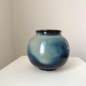May include: A spherical ceramic vase in shades of blue. The vase features a dark blue rim and base, with lighter blue tones in the middle. The glossy surface reflects light, emphasizing its smooth texture. Approximately 15 cm in height.