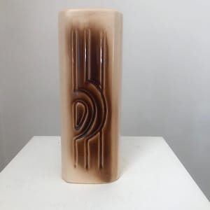 Puede incluir: Un jarrón de cerámica alto y rectangular, con un esmalte marrón claro y un diseño geométrico marrón oscuro. El diseño presenta líneas verticales y una forma curva.