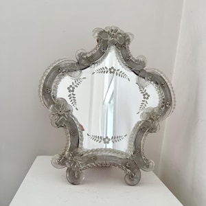 May include: An ornate, clear glass mirror with a distinctive shape. The mirror has a floral etched design around the edges and in the centre. The frame features floral accents and a scalloped edge. The mirror is resting on a white surface.