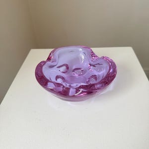 May include: A vintage, lavender-coloured glass ashtray with a wavy rim and a rounded, organic shape. The ashtray has a glossy finish and is displayed on a white surface. The ashtray has several indentations for resting cigarettes.