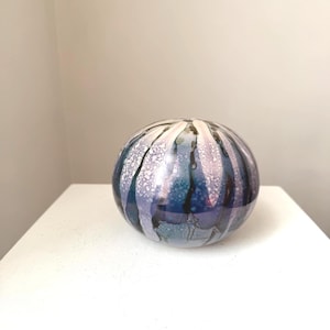 以下が含まれることがあります： 青、紫、白のストライプ模様が施された丸いガラス製の装飾品。 ガラスは光沢仕上げで、白い表面に置かれています。 装飾用の花瓶またはペーパーウェイトのように見えます。