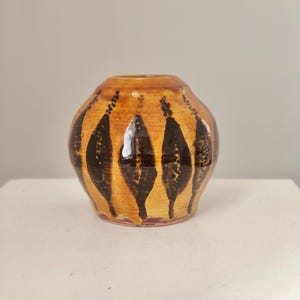 以下が含まれることがあります： 温かみのある黄褐色のベースカラーの、小さくて丸いセラミック花瓶。 垂直の黒い葉のような模様が花瓶の周りに描かれています。 花瓶は白い表面に置かれています。