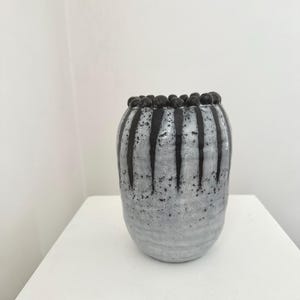 以下が含まれることがあります： 手作りの陶器の花瓶。まだら模様のグレーと白の仕上がり。垂直の黒いストライプが側面を走り、小さな黒いビーズが縁を飾っています。花瓶は白い表面に展示されています。