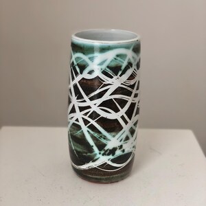 May include: A white ceramic vase with a brown and green abstract design. The vase has a cylindrical shape and a white glaze.