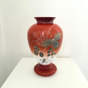 Puede incluir: Un jarrón de cerámica rojo y blanco con un diseño floral. El jarrón tiene un cuerpo bulboso, un borde acampanado y una base redonda. El diseño floral incluye hojas verdes y pequeñas flores blancas.