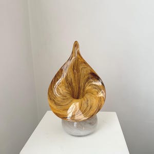 Könnte beinhalten: Eine dekorative Glasskulptur in Tropfenform mit wirbelnden Mustern in Bernstein und Braun. Die Skulptur steht auf einem runden, marmorähnlichen Sockel. Das Kunstwerk wird auf einer weißen Oberfläche ausgestellt.