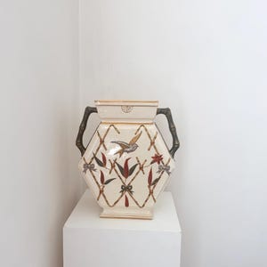 Könnte beinhalten: Cremefarbene Keramikvase mit sechseckigem Körper und Bambusgriffen. Die Vase zeigt ein Vogel- und Blumendesign mit roten, braunen und grünen Akzenten. Das Design ist vor einem Bambusgittermuster angeordnet.