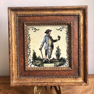May include: A framed artwork featuring a detailed illustration of a man in period clothing holding a shovel and flowers, with the word "Spring" below. The frame is ornate with a gold and brown finish, and the artwork is displayed on a gold easel.