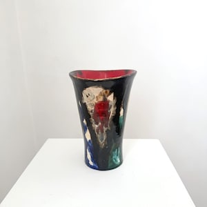 May include: A tall, black ceramic vase with a flared top and abstract painted design. The interior is red, and the exterior features patches of white, blue, and green. The vase is displayed on a white surface.