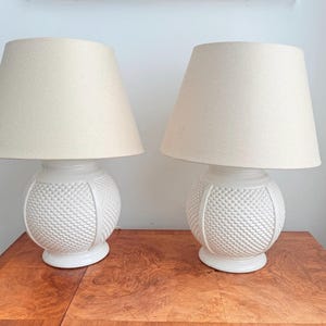 Op de afbeelding: Twee witte keramische tafellampen met getextureerde bolvormige voetstukken en crèmekleurige stoffen kappen. De lampen staan op een houten oppervlak en bieden een klassieke verlichtingsoplossing voor een woon- of slaapkamer.