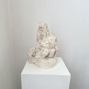 Könnte beinhalten: Eine strukturierte, cremefarbene Skulptur mit abstrakter Form. Die Skulptur steht auf einem weißen Sockel vor einer schlichten weißen Wand. Die Oberfläche der Skulptur weist eine raue, unebene Textur mit verschiedenen Vorsprüngen und Vertiefungen auf.