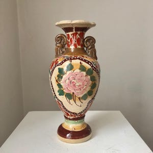 May include: A ceramic vase with a floral design. The vase is decorated with a pink rose, green leaves, and a geometric pattern in red, brown, and white. The vase has two handles on the sides and a flared rim.