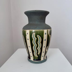 Puede incluir: Un jarrón de cerámica con borde y base verde azulado oscuro. El cuerpo presenta rayas verticales verdes y crema, con puntos azules y detalles ondulados en verde azulado. Es un objeto decorativo.