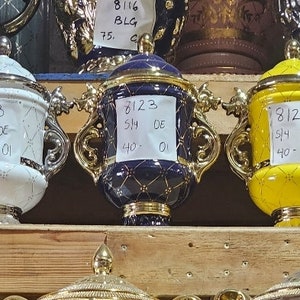 Könnte beinhalten: Eine Regal-Ausstellung mit dekorativen Keramikurnen mit goldenen Akzenten. Die Urnen sind in verschiedenen Farben, darunter Weiß, Gelb, Blau und Braun. Jede Urne hat ein handgeschriebenes Etikett mit Zahlen und Buchstaben.