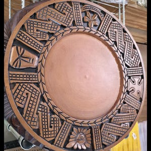 May include: A round, wooden decorative plate with detailed carvings. The plate has a central copper-coloured area surrounded by a border with geometric and floral patterns. The outer edge is dark brown.
