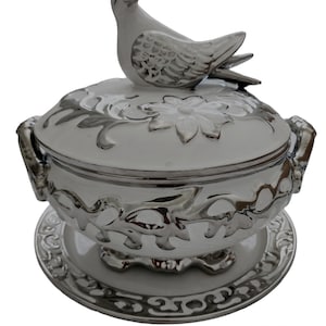 Puede incluir: Tazón de servir de cerámica blanca con un diseño floral plateado y una tapa con forma de pájaro. El tazón tiene un plato a juego.