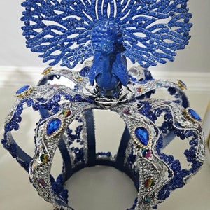 Może przedstawiać: Niebieska korona z srebrnym wykończeniem i wzorem pawia na górze. Korona jest ozdobiona niebieskimi i srebrnymi klejnotami.