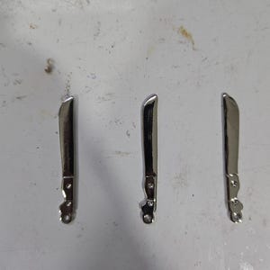 Op de afbeelding: Drie zilverkleurige metalen bedels in de vorm van een machete.