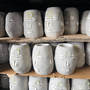 May include: A collection of gray ceramic planters with a face design. Each planter has a circle of small white seashells around the forehead and eyes, and a larger seashell for a nose. Some planters have a green and yellow circle around the forehead.