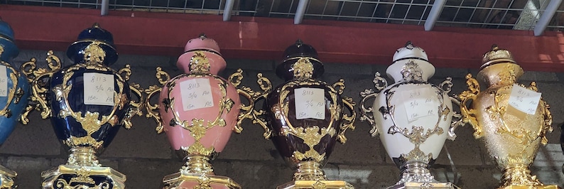 Pode incluir: Seis bules de ch&aacute; decorativos de cer&acirc;mica adornados em v&aacute;rias cores, incluindo azul, rosa, marrom, branco e dourado. Cada bule tem uma tampa dourada e detalhes dourados intrincados. Os bules est&atilde;o dispostos em uma fila em uma prateleira.