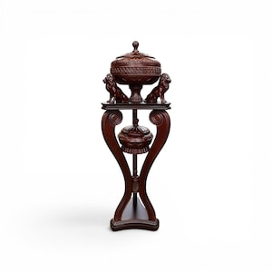 Puede incluir: Un soporte de madera marrón oscuro con base curva y plataforma superior. Dos figuras de leones talladas flanquean un cuenco con tapa en el nivel superior. Un segundo cuenco con tapa más pequeño cuelga en el centro de la estructura.