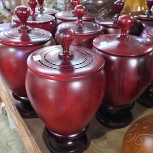 May include: A collection of handcrafted, dark red wooden jars with lids. Each jar has a rounded body, a circular lid, and a small, decorative knob on top. The jars are displayed on a wooden surface, showcasing their rich color and craftsmanship.