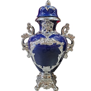 Pode incluir: Um vaso de cer&acirc;mica azul e prata com tampa e detalhes ornamentados. O vaso tem um corpo redondo com uma base larga e duas al&ccedil;as. A tampa &eacute; decorada com um desenho floral.