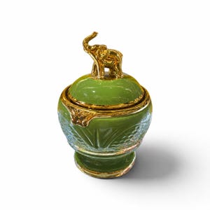 May include: A decorative jar with a clear glass base featuring a textured pattern and a smooth olive green lid. The lid is topped with a metallic gold elephant figurine. The jar has a gold-colored rim where the lid meets the glass base.
