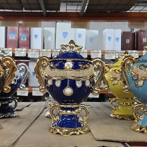 May include: A collection of decorative urns in various colours, including brown, blue, yellow, and light blue, with gold detailing. Each urn features a lid, handles, and jewel and floral embellishments. The urns are displayed on a shelf.