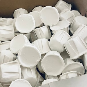 Puede incluir: Primer plano de una caja llena de cápsulas para cupcakes de papel blanco. Las cápsulas están dispuestas en un patrón aleatorio, con algunas de ellas mostrando sus bordes acanalados.