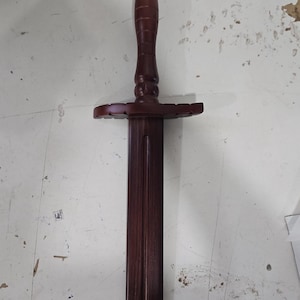 May include: A wooden sword with a dark brown finish. The handle is cylindrical with carved ridges, and the crossguard is a flat, stylized design. The blade is long and rectangular, with a pointed tip. The sword is displayed on a white surface.