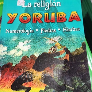 May include: A green book with the title "La religión YORUBA" and the subtitle "Numerología • Piedras • Hierbas". The cover features a colorful illustration of a person wearing a mask and holding a weapon.