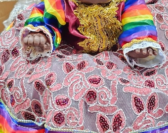 May include: A doll wearing a colourful rainbow patterned dress with a gold crown and a pink and silver floral skirt. The doll has dark skin and black hair.