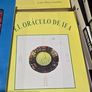 May include: A yellow book with the title "EL ORACULO DE IFA" in green lettering. The book features a black and white image of a circle containing a symbol. The author's name, "Luis Diaz Castillo", is printed in green at the bottom of the cover.