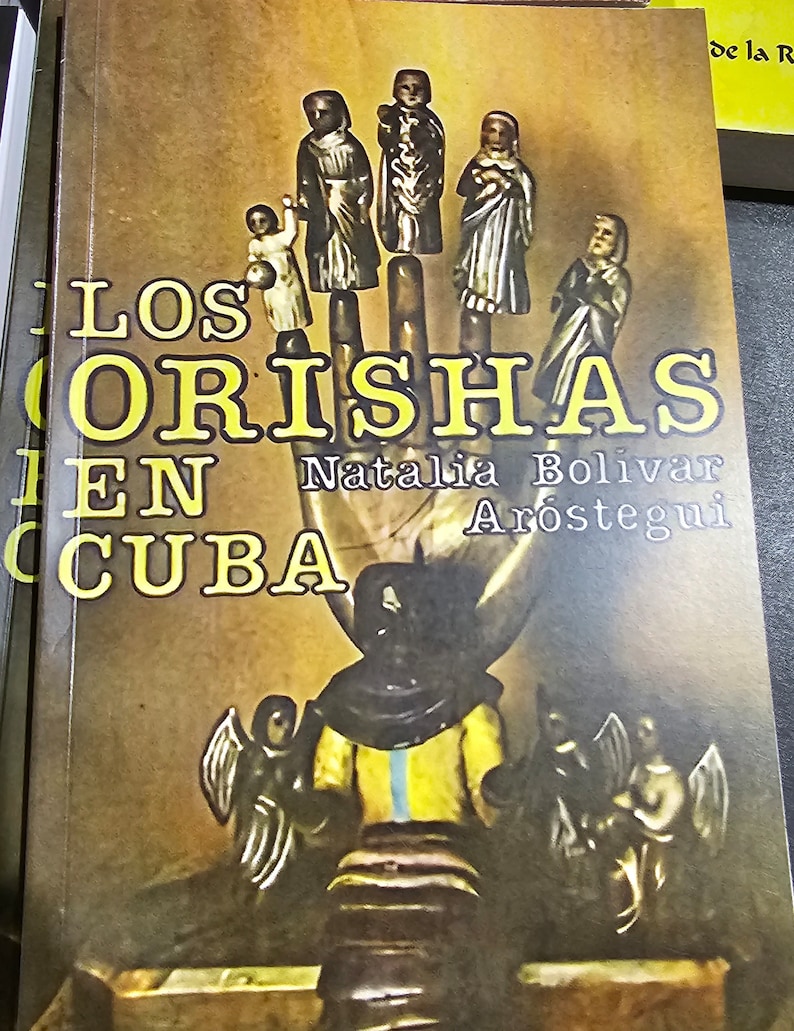 May include: A book cover with a yellow background and the title "Los Orishas en Cuba" in large yellow letters. The cover features a hand with several small figures on the fingers, and a larger figure in the palm of the hand. The author's name, Natalia Bolivar Ar&oacute;stegui, is printed below the title.