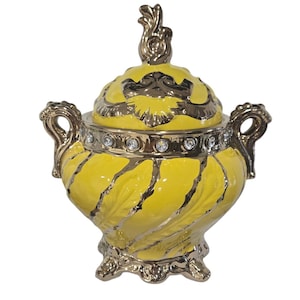 May include: A yellow ceramic serving bowl with a gold lid and decorative accents. The bowl has a swirl pattern and a gold base with ornate feet.