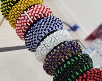 Puede incluir: Una pila de pulseras de cuentas de colores, cada una con un patrón diferente. Las pulseras se muestran en un soporte de madera con una varilla de metal.
