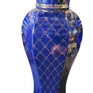 May include: A tall, cobalt blue ceramic vase with a flared top and base. The vase is adorned with a gold geometric pattern and gold trim. The vase has a glossy finish.