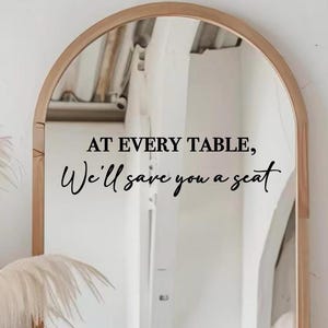 May include: A wooden-framed, arched mirror with the text "AT EVERY TABLE, We'll save you a seat" in black script. The mirror reflects a bright, white interior with architectural details and decorative plants.