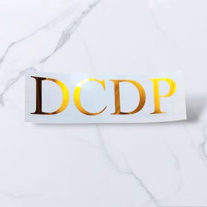 May include: Gold vinyl decal with the letters "DCDP" in a serif font.
