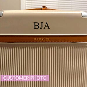 May include: A beige suitcase with a brown leather trim and the word "PARAVEL" embossed on the trim. The suitcase has a personalised monogram "BJA" on the front.