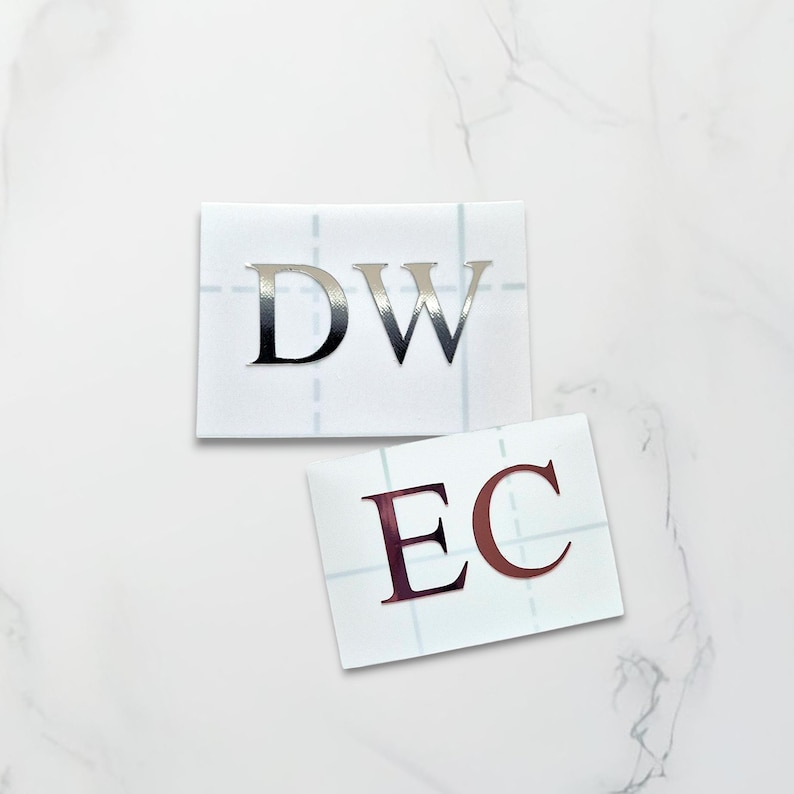 May include: Two sheets of white paper with adhesive letters. The first sheet has the letters "DW" in silver foil. The second sheet has the letters "EC" in rose gold foil.