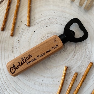 Könnte beinhalten: Holz-Flaschenöffner mit einem schwarzen Metallöffner. Der Griff ist mit dem Text "Christian Bester Papa der Welt" und zwei kleinen Herzen graviert.