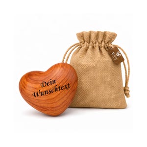 Könnte beinhalten: Ein herzförmiges Objekt aus Holz mit dem Text "Dein Wunschtext" in Schwarz, neben einem Jute-Kordelzugbeutel. Das Herz hat eine natürliche Holzmaserung. Der Beutel hat ein kleines Etikett.