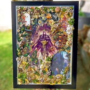 Può includere: Opera d'arte incorniciata a tecnica mista con una figura centrale in mantello viola, circondata da elementi floreali, farfalle e un'illustrazione di un castello. L'opera è in una cornice nera, con sfondo di fogliame verde.