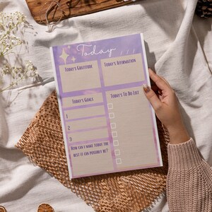 May include: A pink and purple daily planner with a watercolor background. The planner has sections for today's gratitude, affirmation, goals, and to-do list. It also includes a section for how to make today the best it can possibly be.