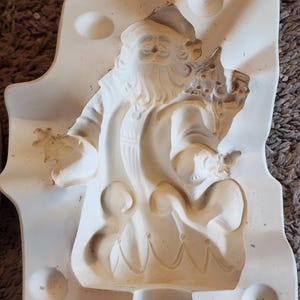 May include: A white ceramic mold in the shape of Santa Claus, holding a sack and a small tree. The mold is in a rectangular shape with rounded edges and circular indentations.