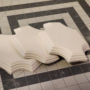May include: Three white, decorative, architectural elements with a curved, layered design. The pieces are arranged on a gray and black grid surface, suggesting a crafting or design setting. The shapes have a smooth, matte finish.