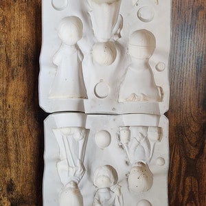 May include: A two-piece white ceramic mold for making figurines. The mold has six cavities, each shaped like a person. The mold is made of a smooth, white material.