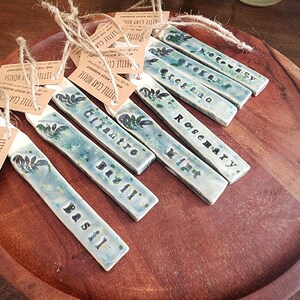 May include: A collection of handmade ceramic garden markers with the names of herbs like basil, cilantro, and rosemary. Each marker is rectangular, with a blue-green glaze and a botanical design. They are attached to tags with twine.